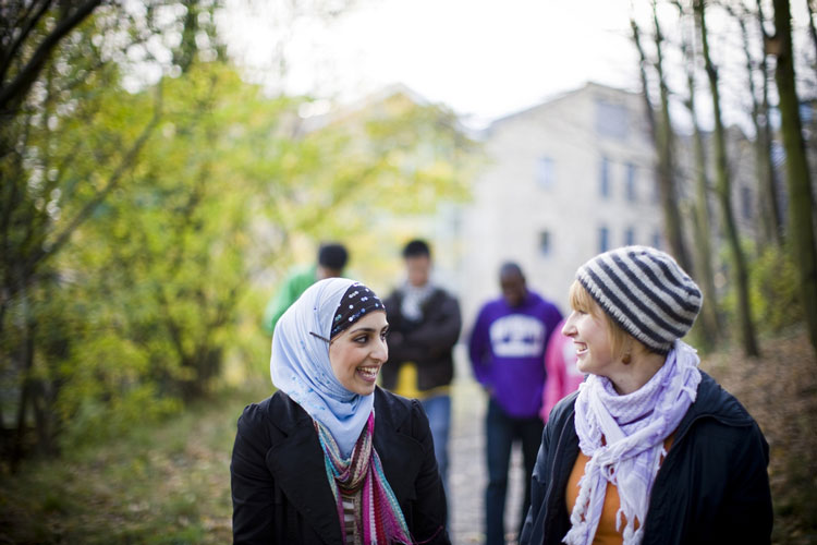 In focus, at the front, two students talk as 3 hang back, out of focus, There are trees to the left and right of the frame and a building in the background.